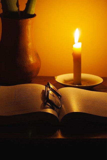 scrivania, con libro aperto, occhiali nel mezzo e sullo sfondo una candela accesa che illumina il buio della stanza